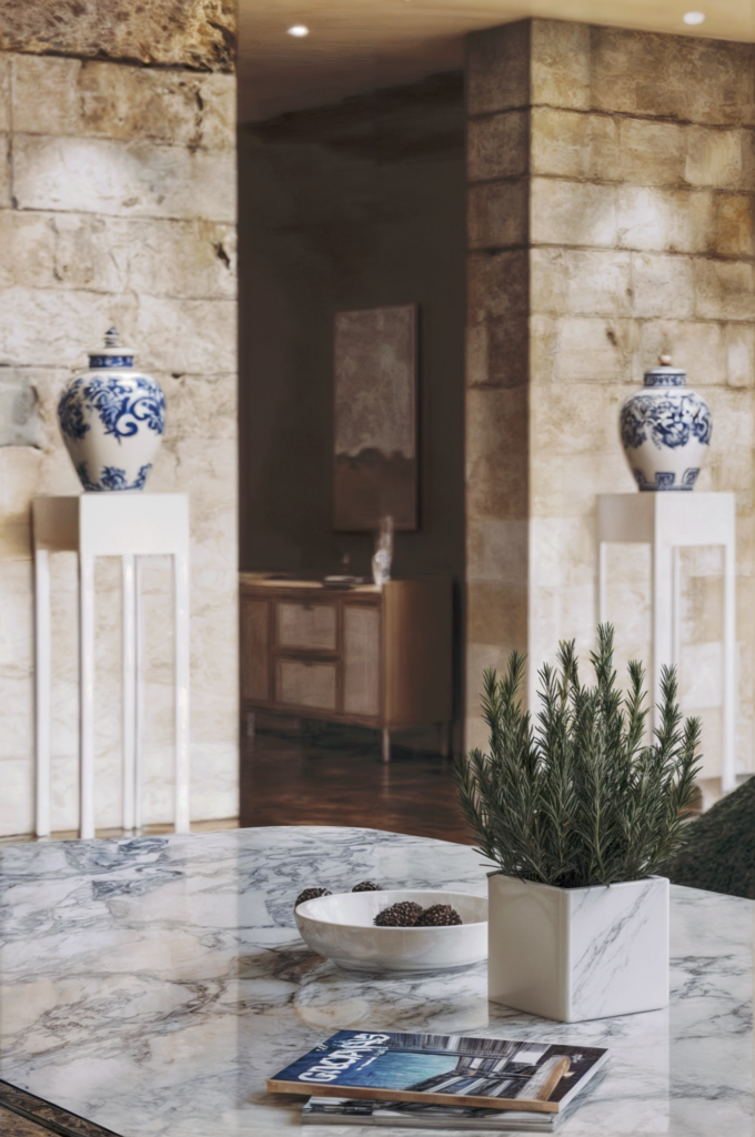 Image du salon après la rénovation par BYAA Inside à Bordeaux Pey-Berland. On y voit une grande ouverture sur l'extérieur, un mur en pierre, un canapé gris, des fauteuils en tissu et une table basse en marbre.